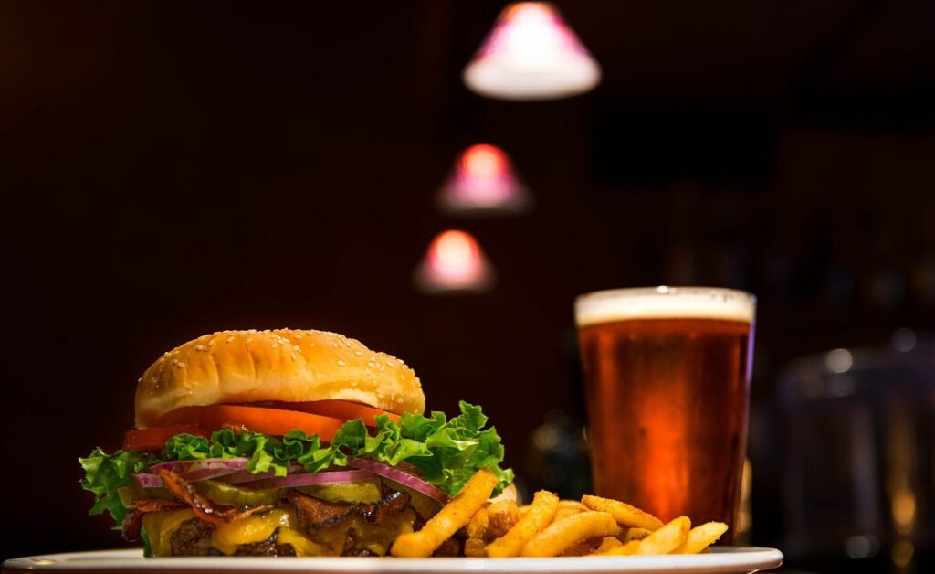 Bacon cheeseburger with seasoned fries and a pint glass on a dark bar table