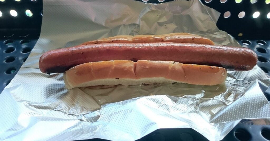 Long grilled hot dog in a bun on foil over a perforated tray